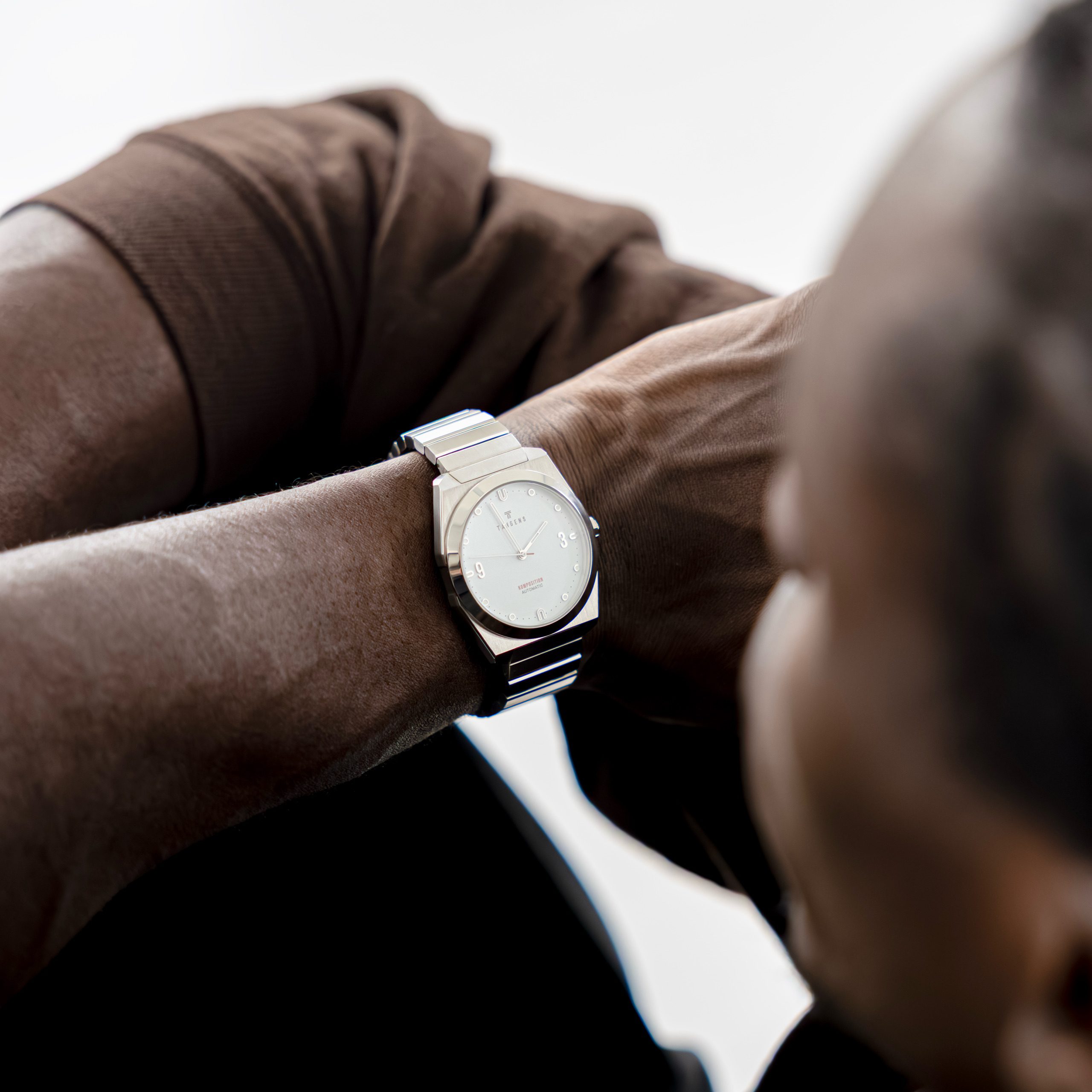 A man in a brown sweater holds his arm across his face, a Komposition Automatic Bright White on his wrist
