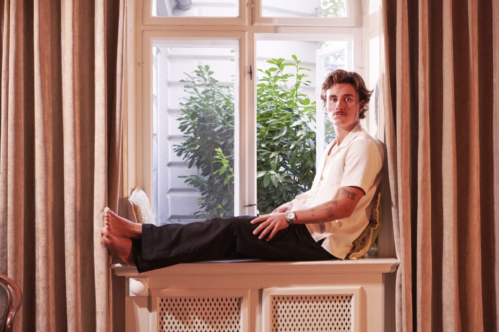 A man sits barefoot on a windowsill between long curtains